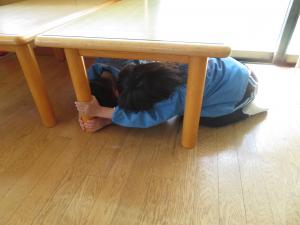 机の下に隠れて、身を守る練習をしているところ