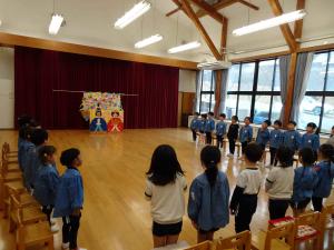 全園児が遊戯室に集まり椅子の前に立っている写真